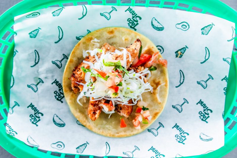 Grilled chicken taco topped with shredded cabbage, diced tomatoes, and herbs on a warm corn tortilla in a green basket.