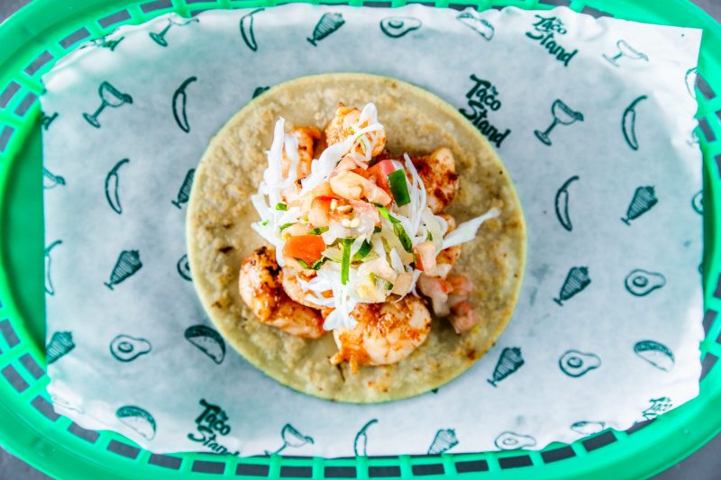 Shrimp taco topped with shredded cabbage slaw and diced vegetables on a corn tortilla in a green basket lined with printed parchmentpaper-themed wrap.