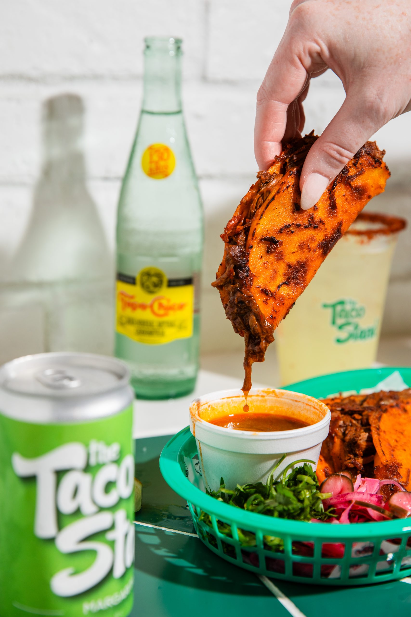 Hand holds a crispy orange-coated taco over a cup of orange sauce, with drink bottles in the blurred background.