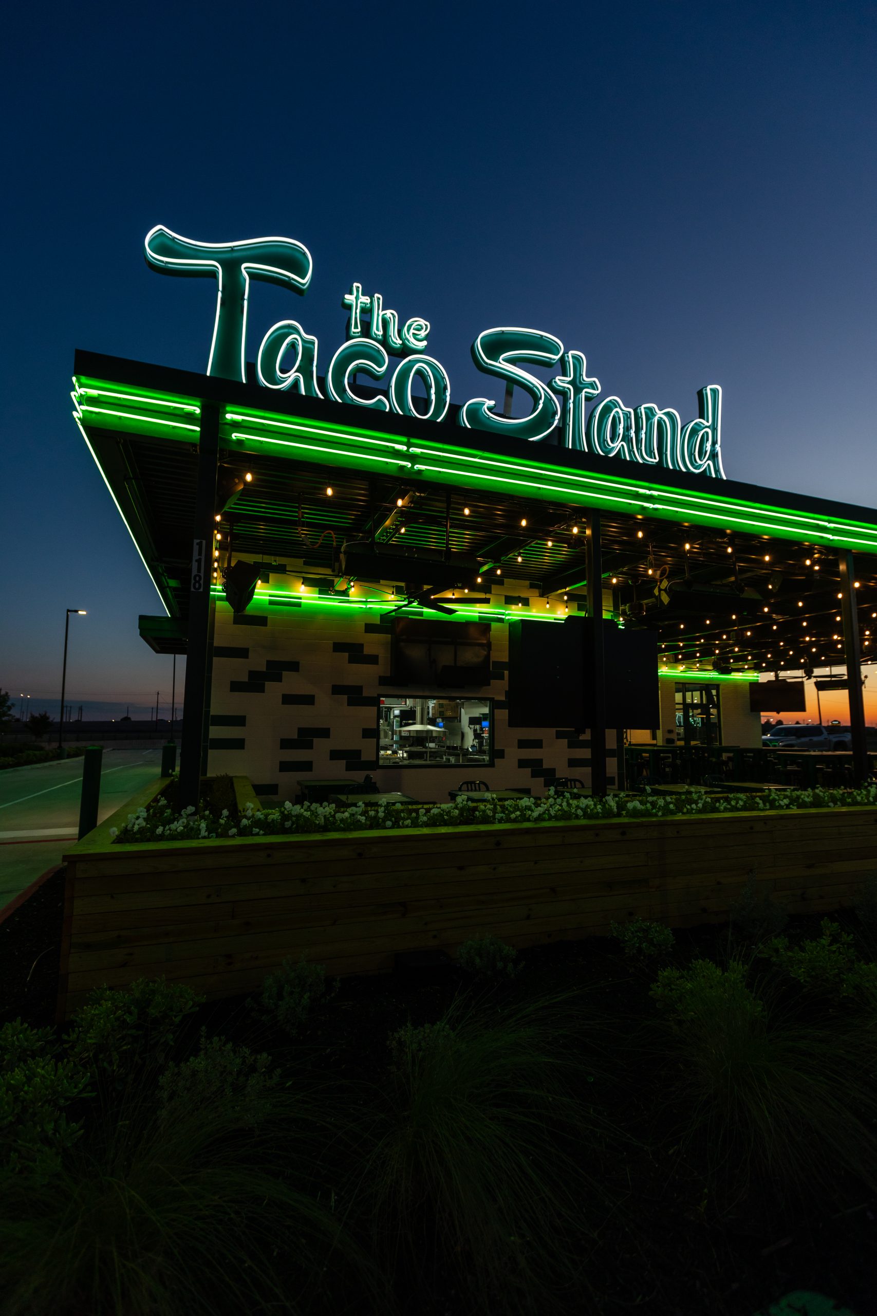 Neon sign reading 'The Taco Stand' above a modern restaurant at dusk with green lighting accents a storefront exterior.