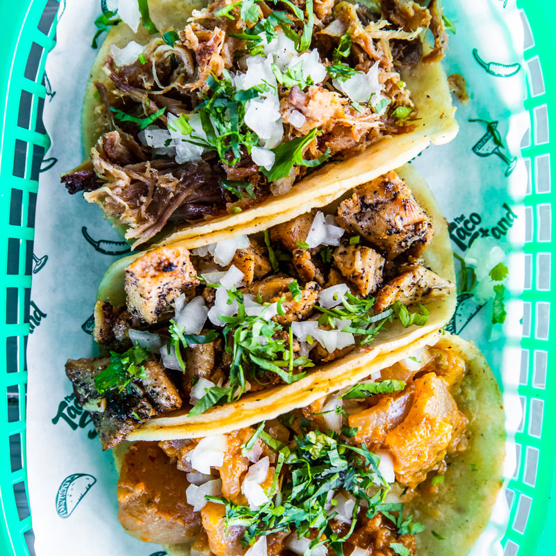 Three street-style tacos filled with sautéed meat, onions, and cilantro in soft corn tortillas, served in a bright green basket with salsa verde on the side (Mexican street-food vibe).